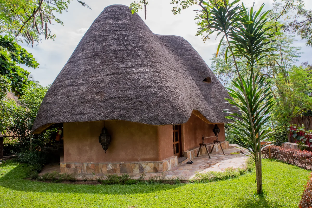 A round thatched bungalow with mud-rendered walls and a stone base, set on a lawn among tropical palms