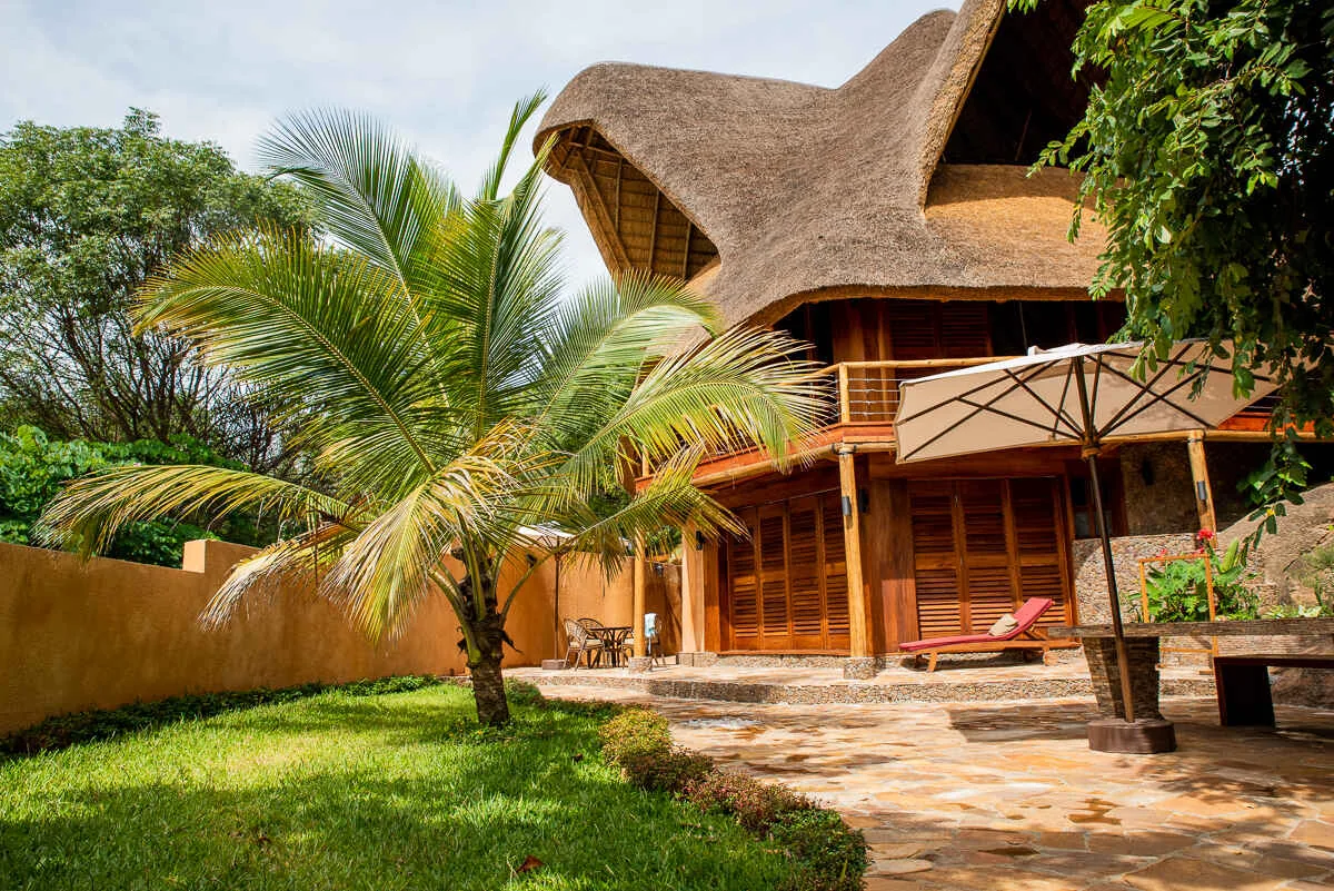 A two-storey thatched villa with a wooden balcony, sun lounger and parasol on its garden terrace