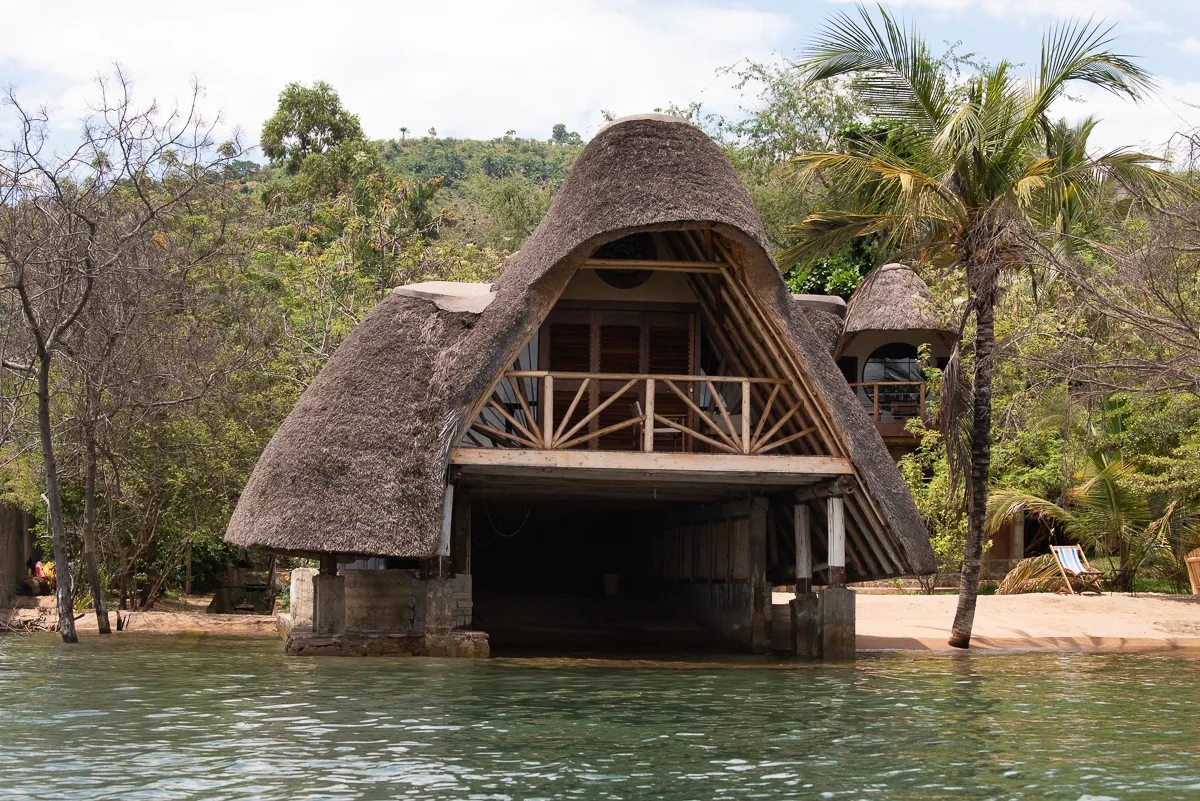 A thatched lakefront building with a railed balcony reaching out over the water, seen from the lake