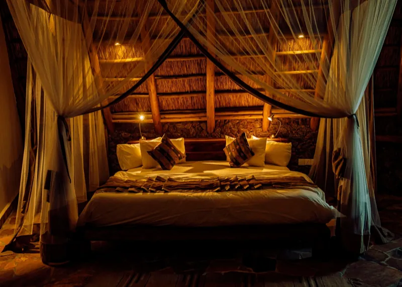 A king bed draped with a mosquito net under a thatched roof, lit by warm lamplight